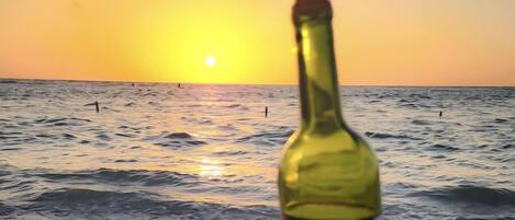 Una botella de vino en la playa con el océano y una puesta de sol al fondo.