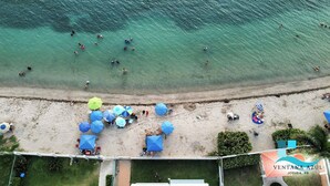 An aerial view of a beach with clear turquoise water, people swimming and sunbathing, and several umbrellas on the sand.
