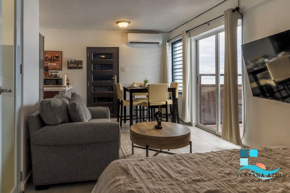 A compact living space with a sofa, a round coffee table, a dining area with chairs, a TV mounted on the wall, and a large window with curtains.