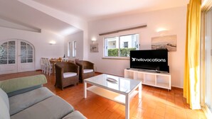 A spacious living room with a glass coffee table, wicker chairs, a sofa, a television, and a dining area with chairs and a table. - Villa Uno by HelloVacations (Loulé)