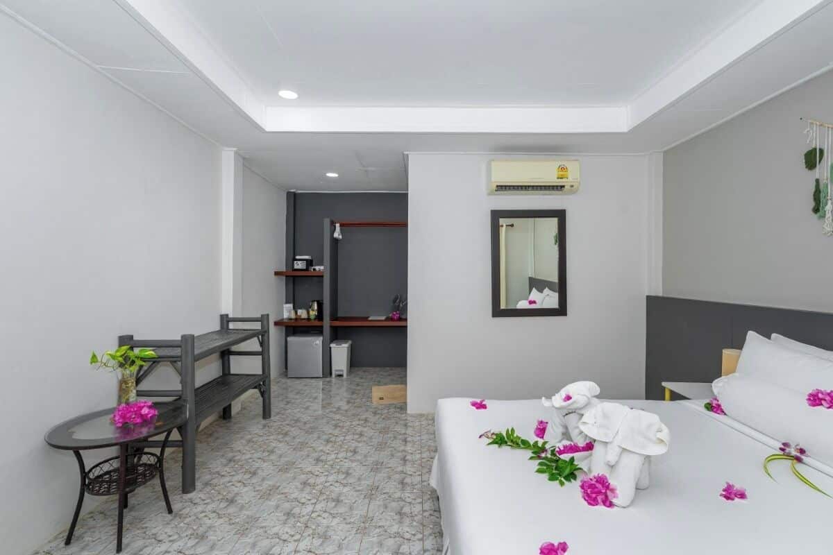 Quarto de hotel com uma cama, uma escrivaninha, uma cadeira, uma mesa pequena com um vaso de flores, um espelho na parede e uma pequena cozinha no fundo.