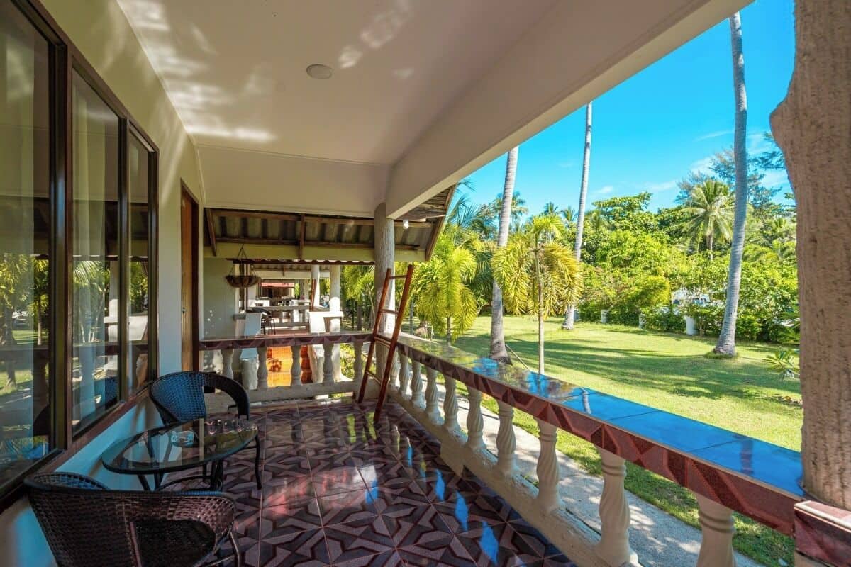 Uma área externa coberta com mesa e cadeiras de vidro, vista para um jardim exuberante e uma sala de jantar ao fundo.