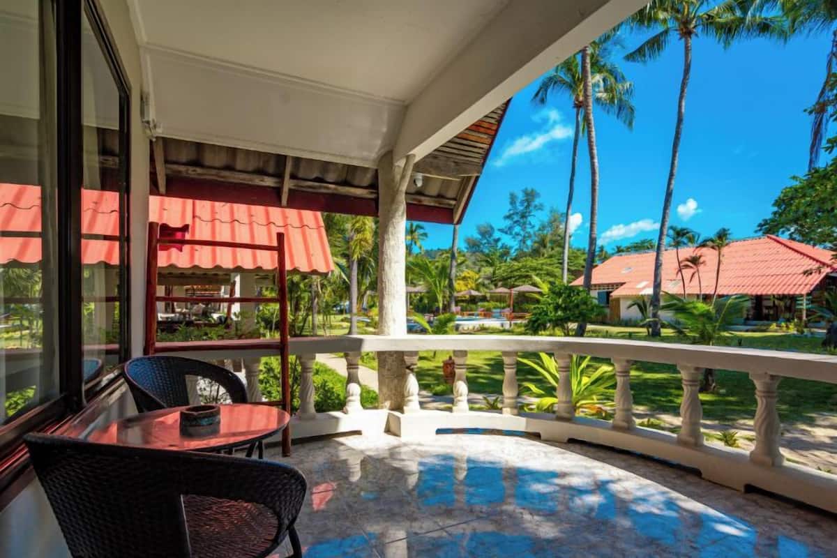 Área externa coberta com mesa, cadeiras, corrimão e vista para uma piscina e palmeiras.