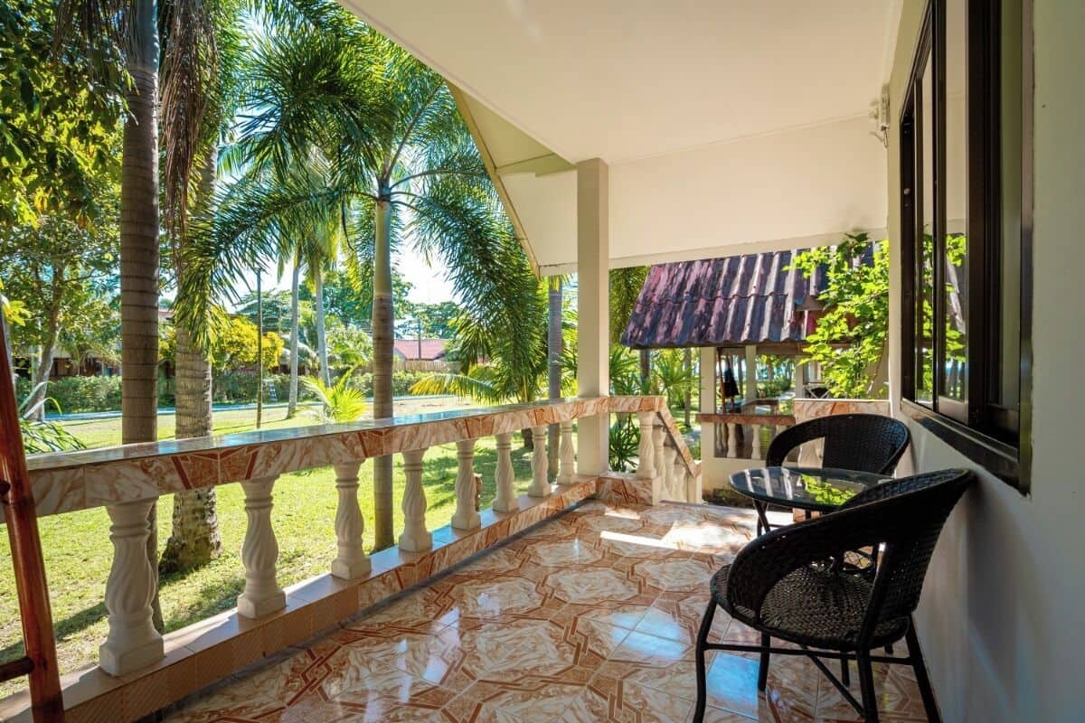 Uma área externa coberta com piso de azulejos, guarda-corpo curvo e vista para um jardim com palmeiras.