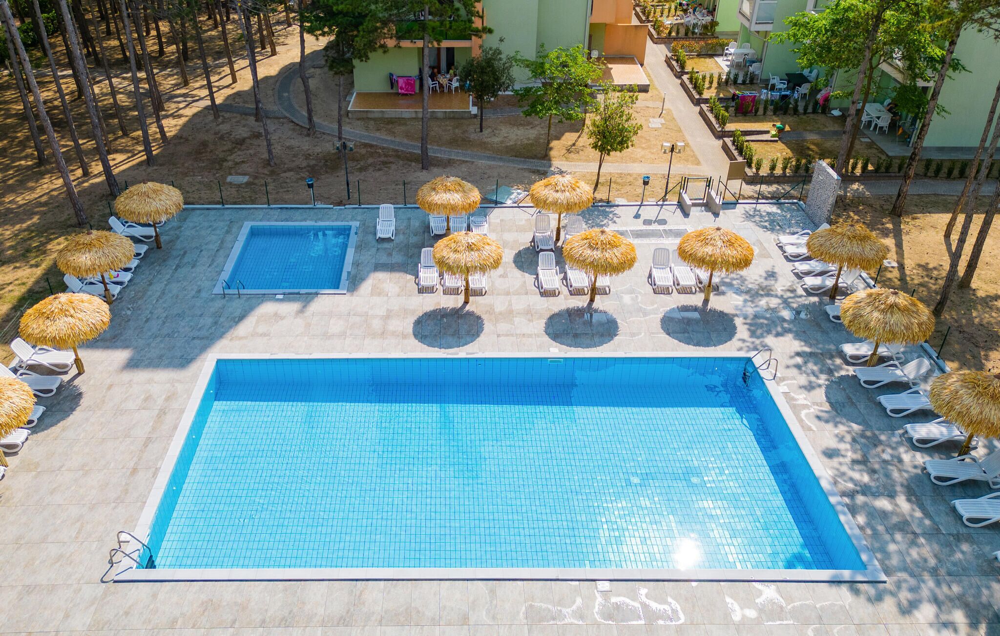 A large rectangular swimming pool with a smaller adjacent pool, surrounded by sun loungers and thatched umbrellas, with a landscaped area and buildings in the background.