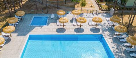 A large rectangular swimming pool with a smaller adjacent pool, surrounded by sun loungers and thatched umbrellas, with a landscaped area and buildings in the background.