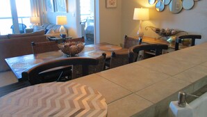 Un comedor con una mesa larga, sillas y un cuenco decorativo. Se observa un sofá y una lámpara al fondo.