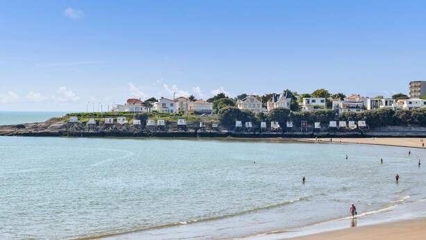 A coastal area with a sandy beach, clear blue water, and a row of houses along the shore.