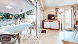 A room with a glass door leading to a patio, a dining table, chairs, a TV, and a small kitchen area.