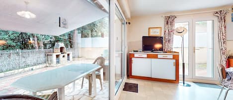 A room with a glass door leading to a patio, a dining table, chairs, a TV, and a small kitchen area.