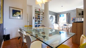 Uma sala de estar moderna com uma mesa de jantar de vidro, cadeiras brancas e amarelas, uma estante e uma janela grande com cortinas.