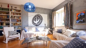 Uma sala de estar com sofá, poltronas, mesa de centro e estante de livros. Há uma janela grande com cortinas, uma luminária pendente azul e um letreiro de boas-vindas na parede.