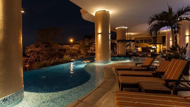 A pool area with lounge chairs and palm trees, surrounded by large pillars and a night sky. - Signature by Pinnacle (Puerto Vallarta)