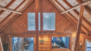 Interior de una cabaña de troncos con techo alto, vigas de madera y ventanas amplias.