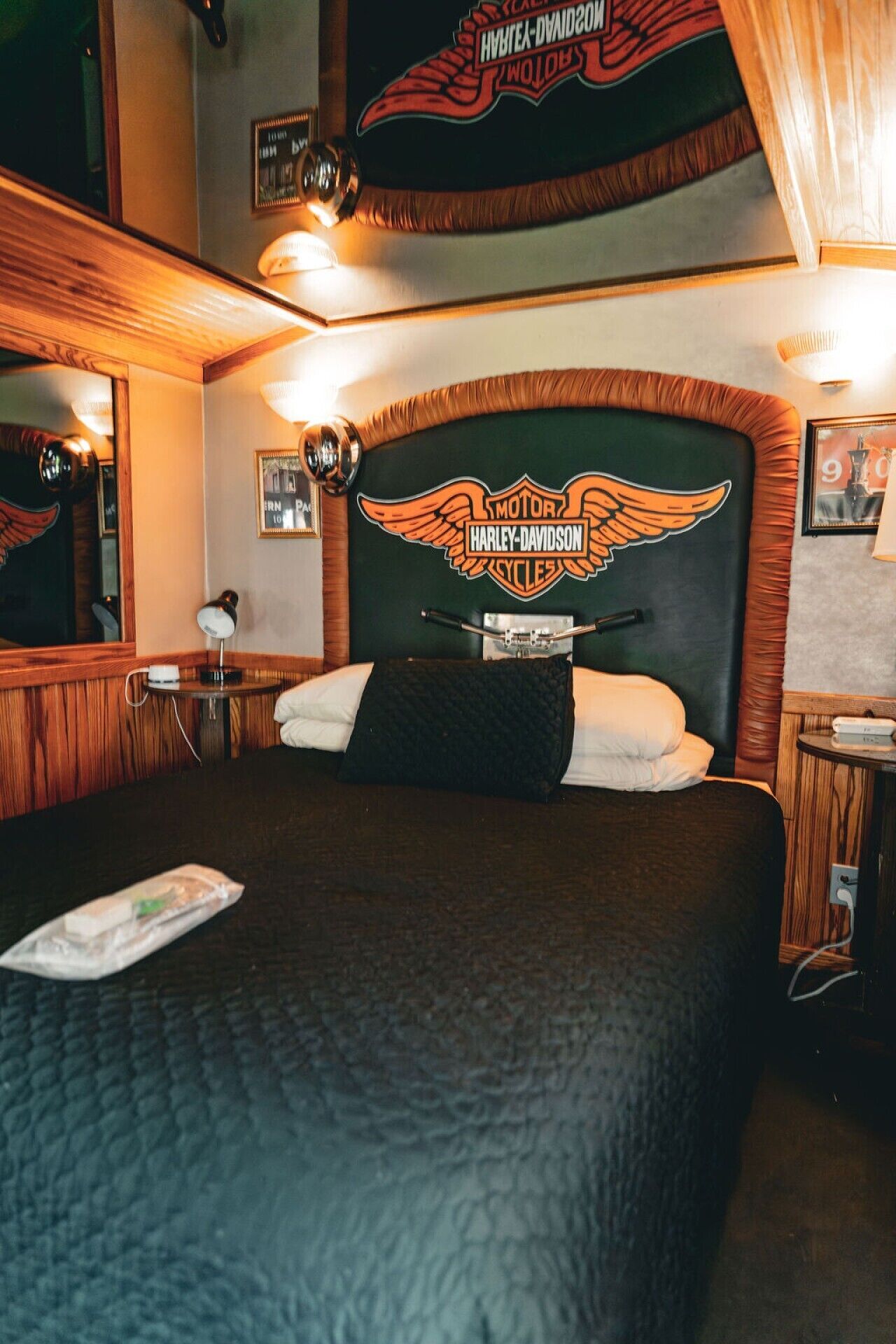 A hotel room with a Harley-Davidson themed headboard, a bed with white and dark bedding, a bedside table with a lamp, and a mirror on the wall.