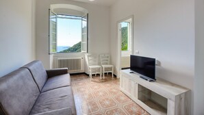A modern living room with a grey sofa, a TV on a wooden stand, two white chairs, and large windows with a view of greenery and the sea. - 2 bedroom gorgeous apartment in Semorile (Semorile)