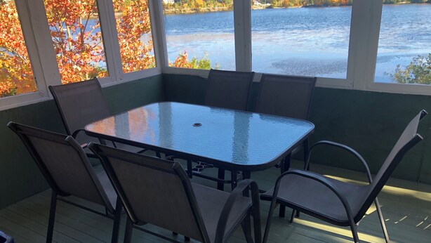 Un espace extérieur abrité, doté d’une table à plateau de verre et de chaises en métal, offrant une vue sur un plan d’eau et le feuillage automnal.