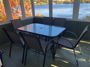 Ein überdachter Außenbereich mit Tischplatte aus Glas und Metallstühlen, mit Blick auf ein Gewässer und Herbstlaub.