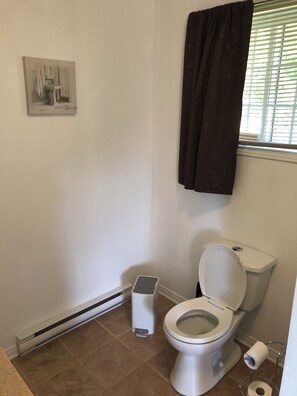 A bathroom with a toilet, a trash can, a radiator, and a window with blinds.