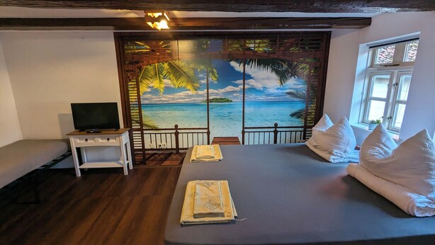 Een slaapkamer met een bed, kussens, een televisie op een klein tafeltje en uitzicht op de oceaan door een groot raam.