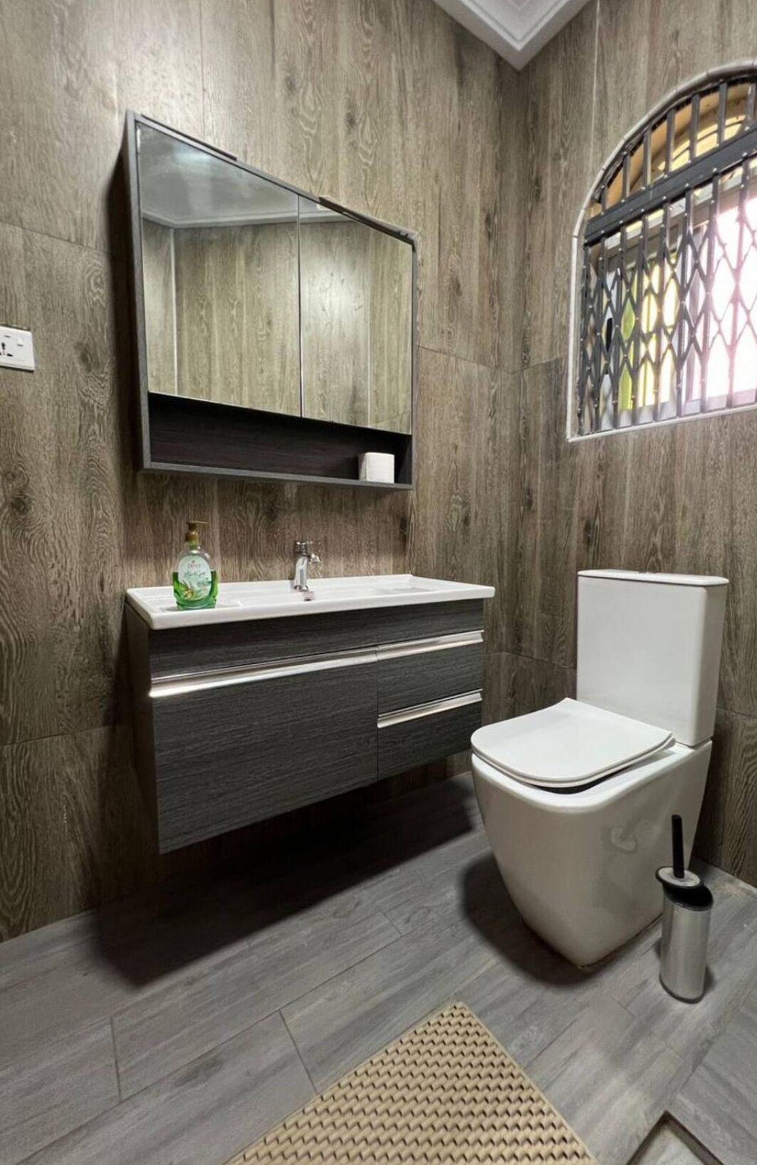 A bathroom with a wooden wall, a white sink, a toilet, and a window with a decorative grille.