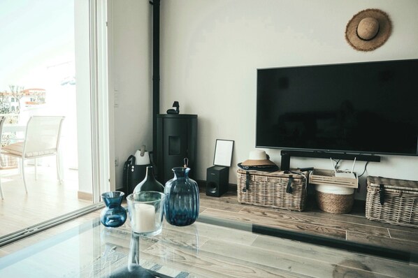 Una sala moderna con una mesa de centro de vidrio, un televisor montado en la pared, cestas de mimbre y un ventanal con vistas al exterior.