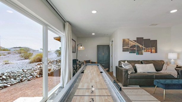 A room with a shuffleboard table, a sofa, and a view of a rocky landscape through large windows. - Havasu Cotati - Pool, Spa, FirePit, Games, Garage (Lake Havasu City)