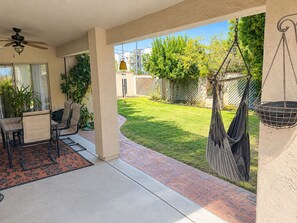 A patio with a table and chairs, a hanging hammock, and a view of a grassy yard with trees.