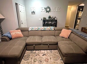 A living room with a sectional sofa, a patterned throw, a 'HOME' sign, and a bookshelf.