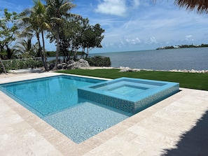 Una piscina rectangular con borde de azulejos, rodeada por un suelo de piedra y una zona ajardinada con palmeras.
