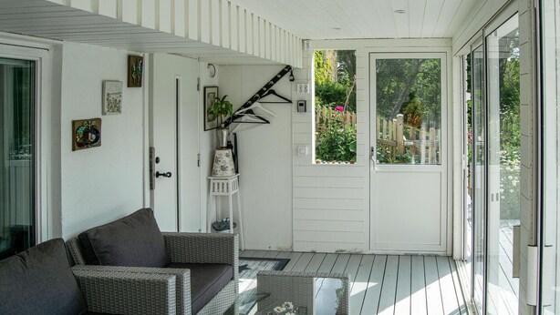 A covered porch with a wicker sofa, a small table with a vase, and a door leading to a garden.