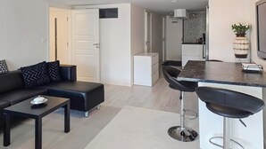 A modern living room with a black leather sofa, a small coffee table, and a dining area with a bar stool and a kitchen in the background.