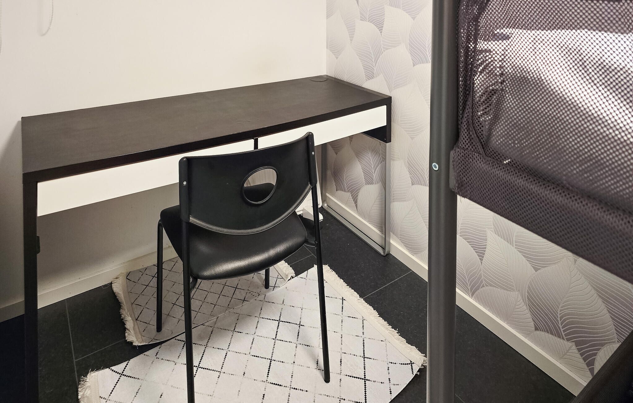 A small, dark-colored desk with a chair in front, a patterned rug, and a mesh screen door.