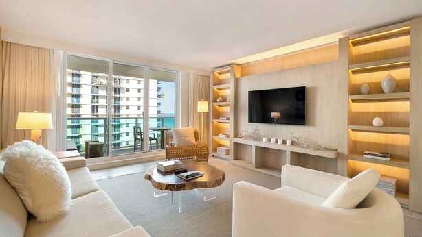 A modern hotel room with a large balcony, a flat-screen TV, a wooden bookshelf, and a comfortable seating area.