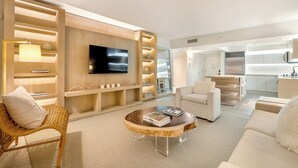 A modern living room with a flat-screen TV, built-in shelving, a coffee table, and a kitchen area in the background.