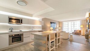 A modern kitchen with integrated appliances, a dining area with a bar, and a living space with a sofa and a large window.