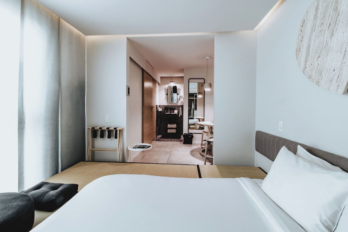 A modern hotel room with a large bed, a small table, and a view into the kitchen area.