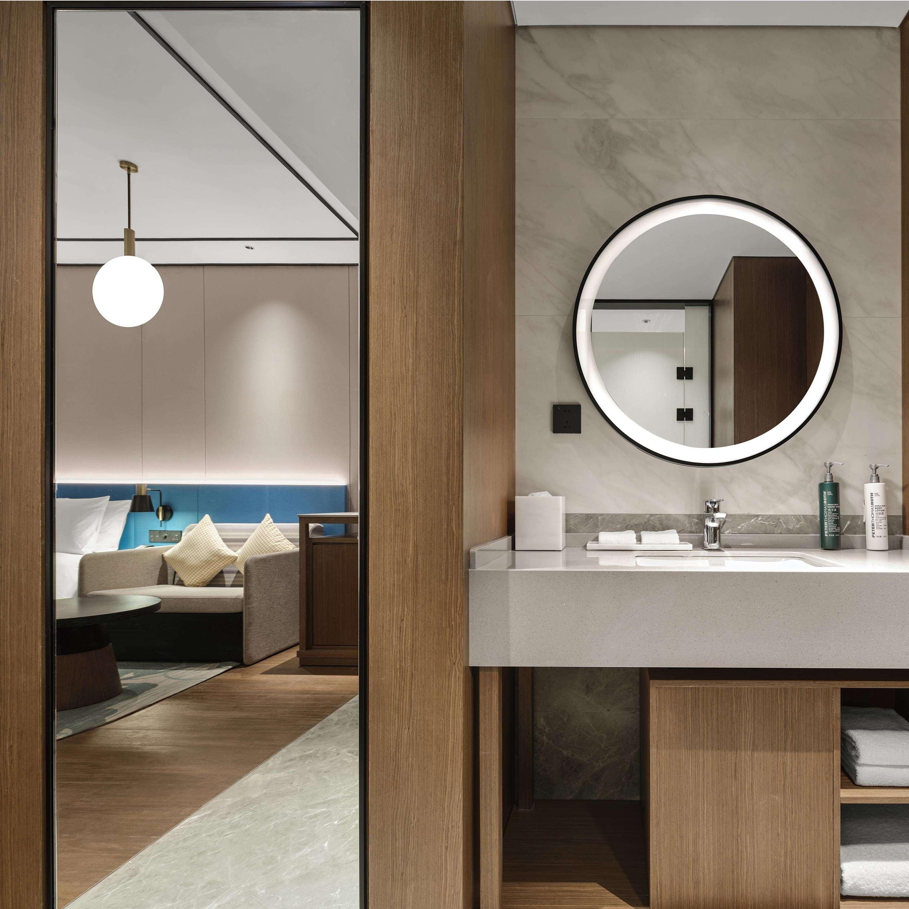 Quarto de hotel moderno com piso de madeira, espelho redondo com moldura preta, pia branca e cama com cabeceira azul.