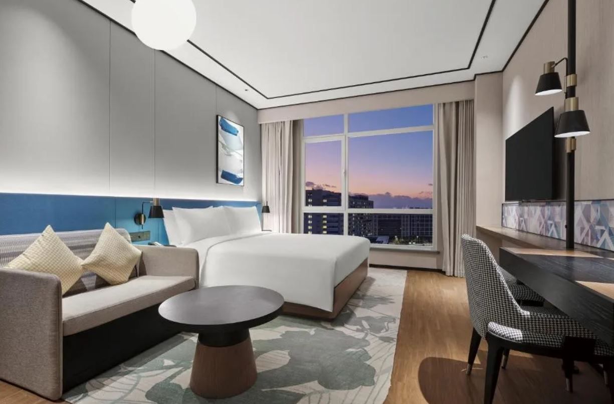 Quarto de hotel moderno com uma cama grande, um sofá, uma mesa e vista da cidade ao pôr do sol.