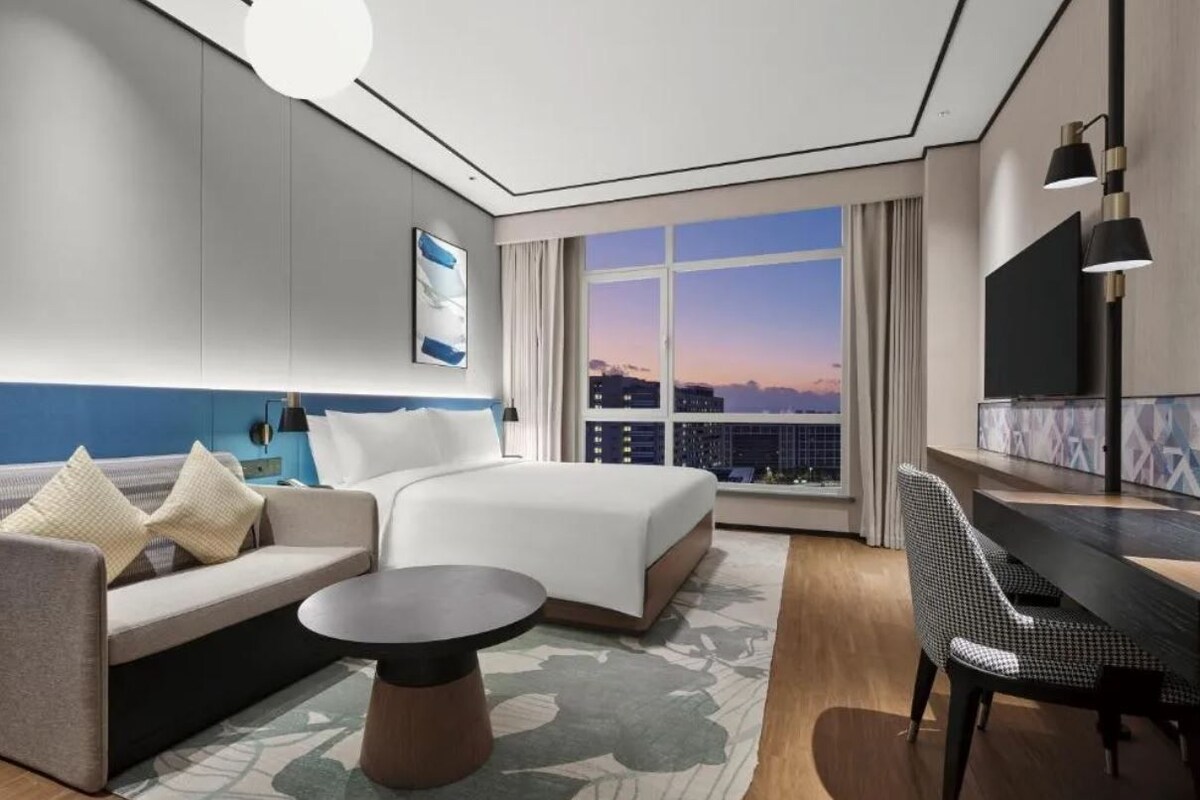 A modern hotel room with a large bed, a sofa, a desk, and a view of the city at sunset.