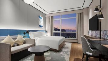 Quarto de hotel moderno com uma cama grande, um sofá, uma mesa e vista da cidade ao pôr do sol.