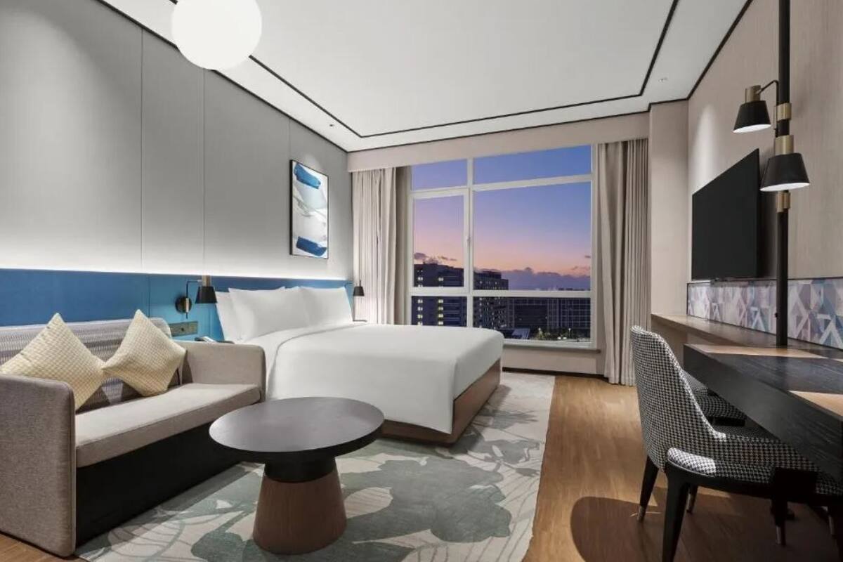 Une chambre d’hôtel moderne, dotée d’un grand lit, d’un canapé, d’un bureau et offrant une vue sur la ville au coucher du soleil.