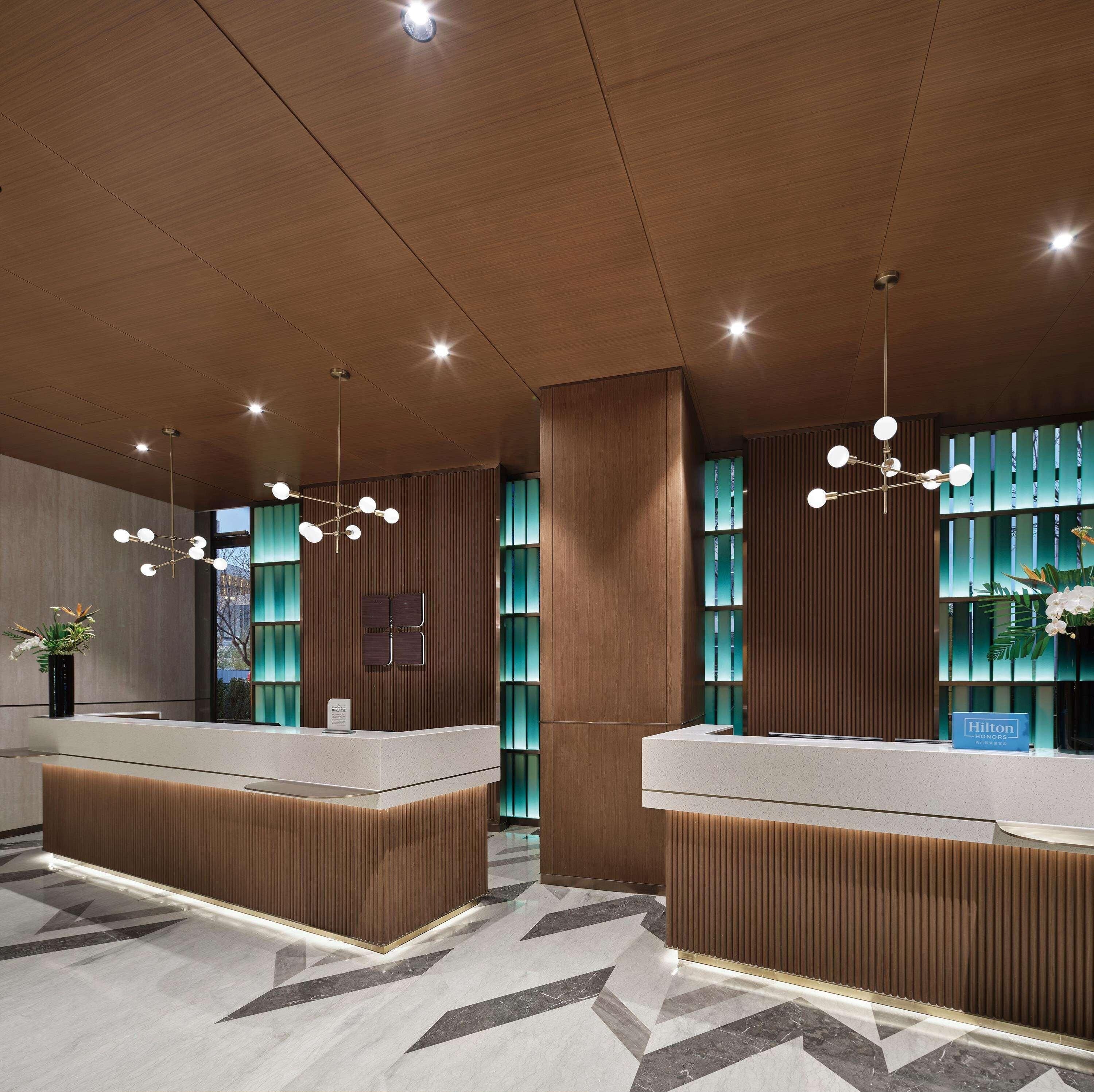 Uma área de recepção de hotel moderna, com painéis de madeira, dois balcões de atendimento, uma grande planta em vaso e um lustre decorativo.