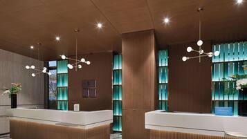 Uma área de recepção de hotel moderna, com painéis de madeira, dois balcões de atendimento, uma grande planta em vaso e um lustre decorativo.
