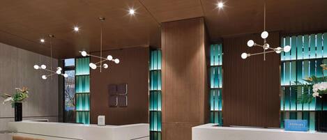 Un área de recepción de hotel moderna con paneles de madera, dos mostradores de recepción, una planta grande en maceta y una lámpara decorativa.