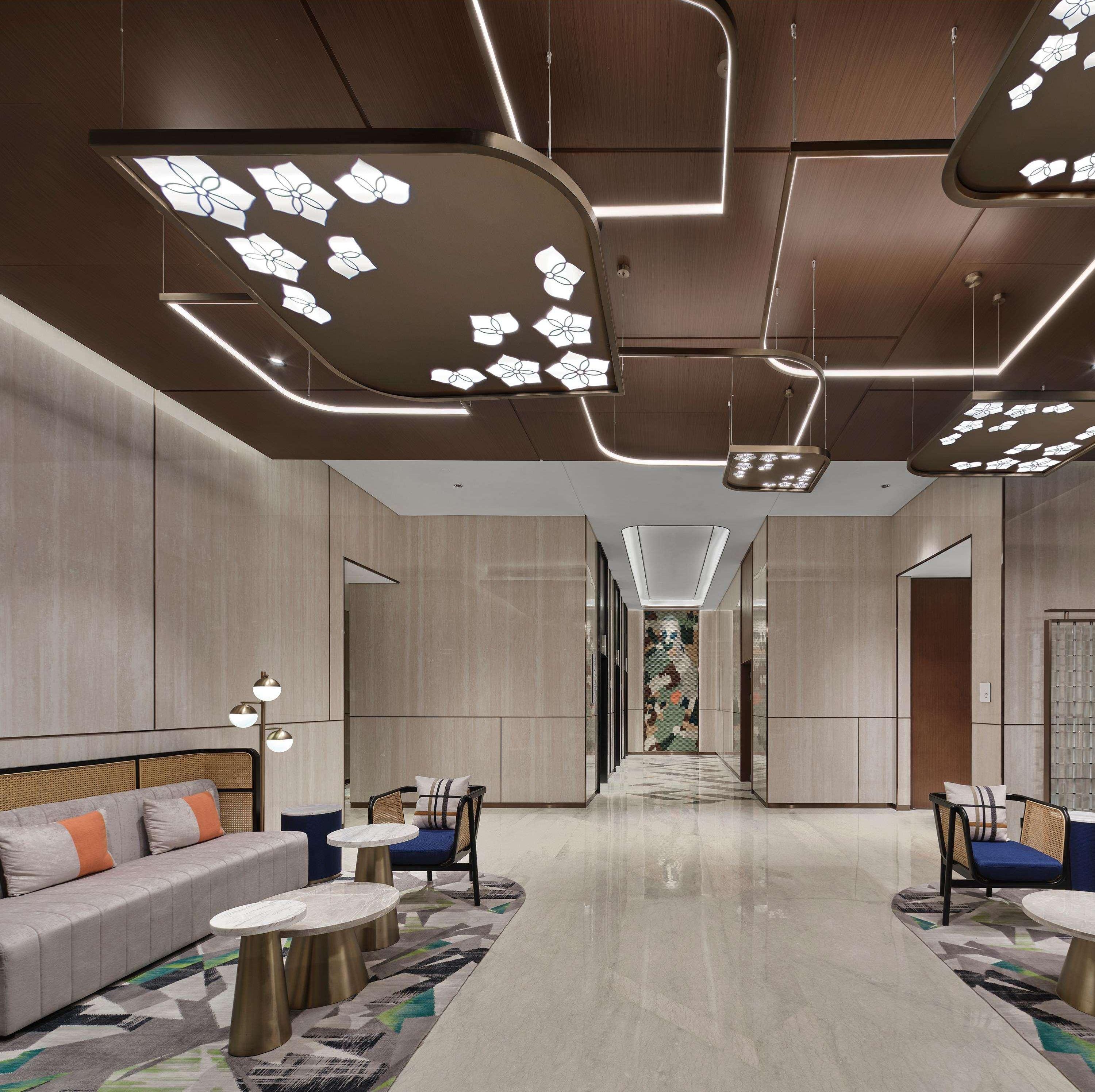 Um hall de hotel moderno com área de estar central, parede estampada e um corredor longo que dá acesso a outros quartos.