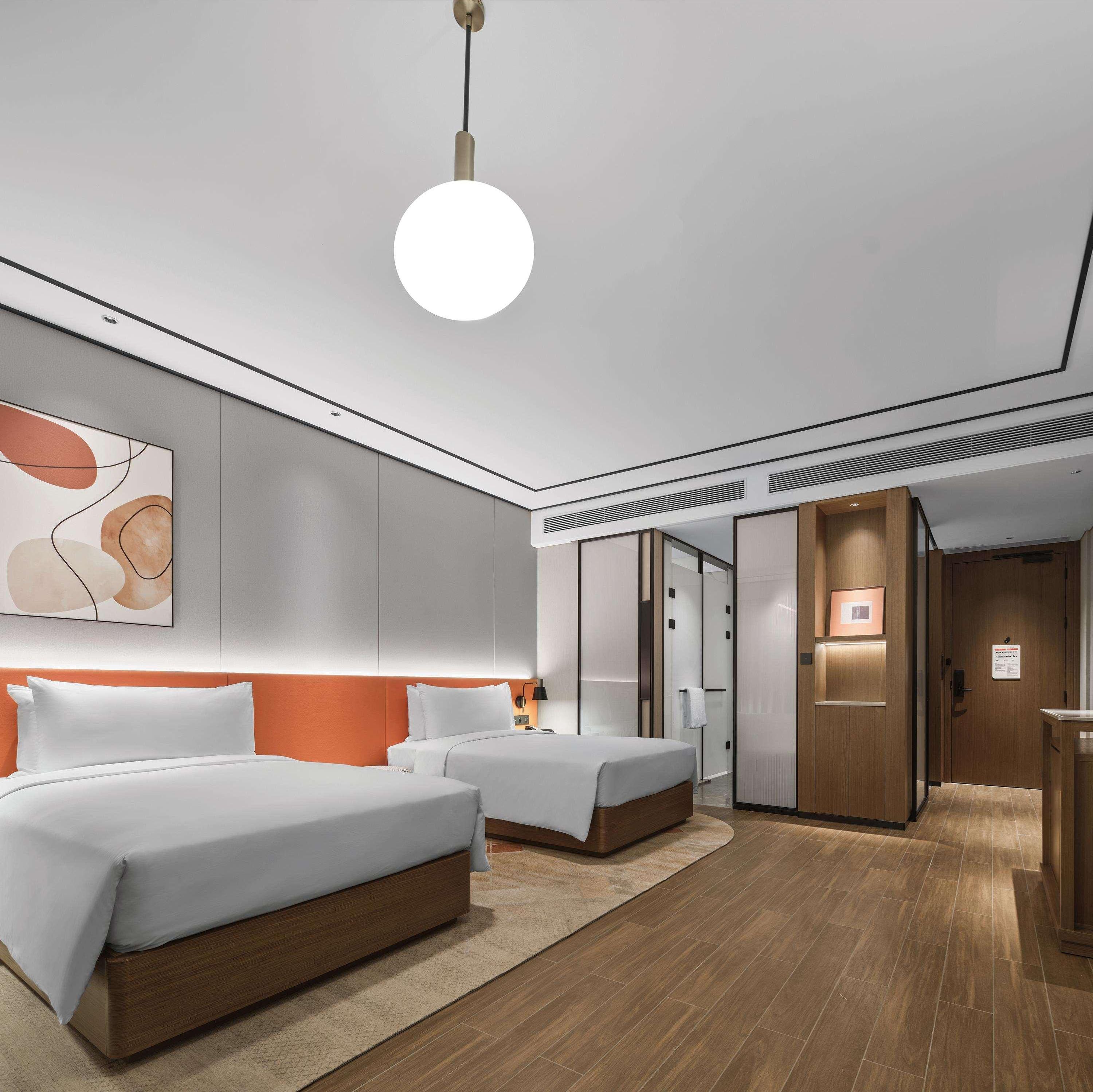 Quarto de hotel moderno com duas camas, uma pintura abstrata grande, uma luminária de teto redonda e piso de madeira.