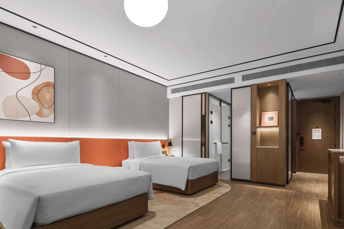 A modern hotel room with two beds, a large abstract painting, a round ceiling light, and a wooden floor.