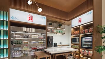 A modern retail space with a central counter, refrigerated beverage display, and shelves stocked with snacks and packaged goods.