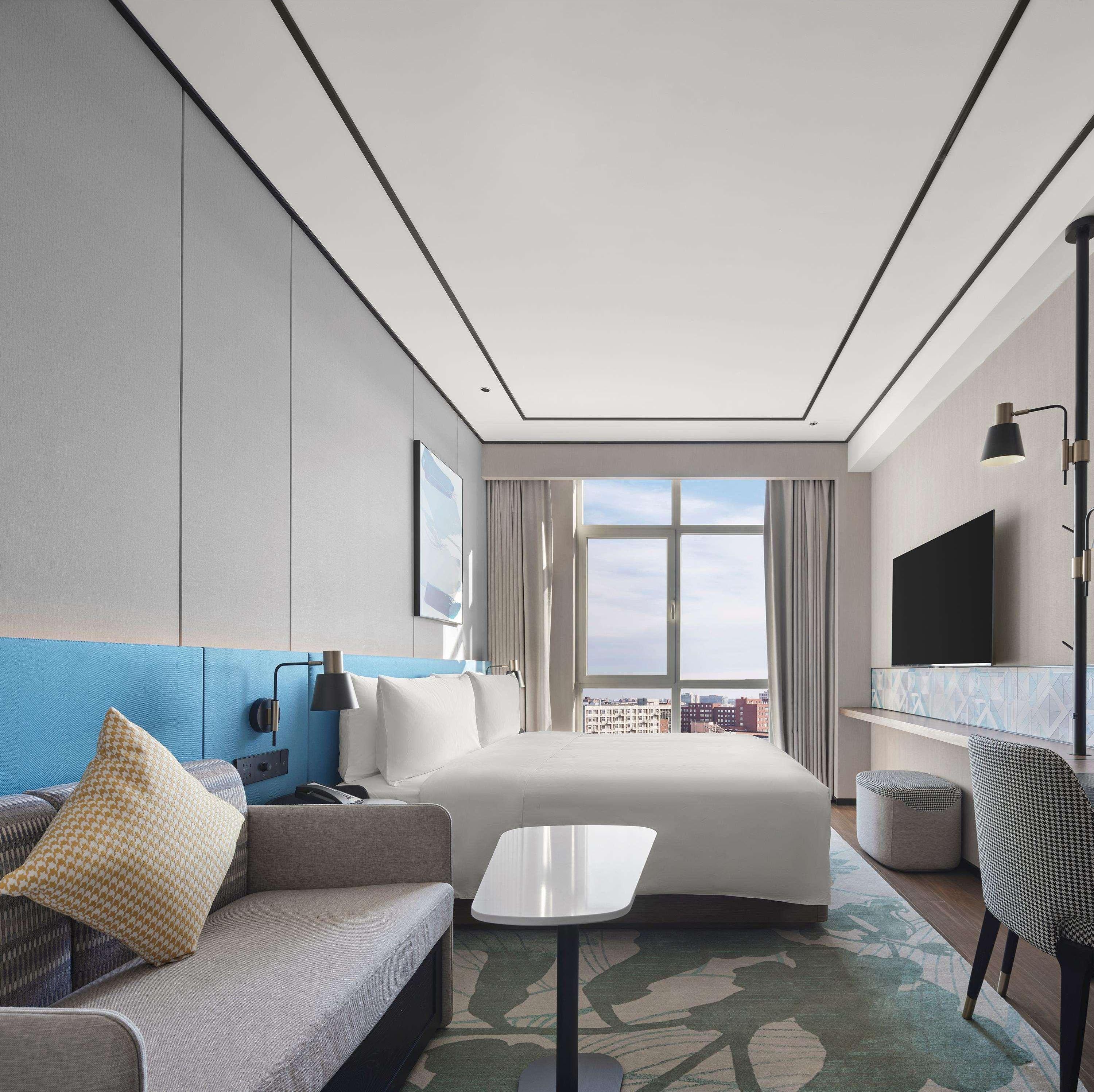 A modern hotel room with a bed, a sofa, a small table, a TV, and a window with a city view.